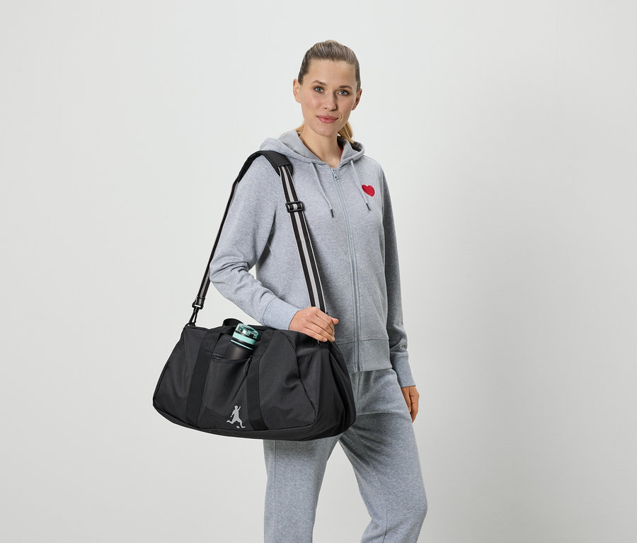 Frau mit grauer Trainingsjacke mit rotem Herz und grauer Trainingshose hält eine schwarze Sporttasche mit Trinkflasche.