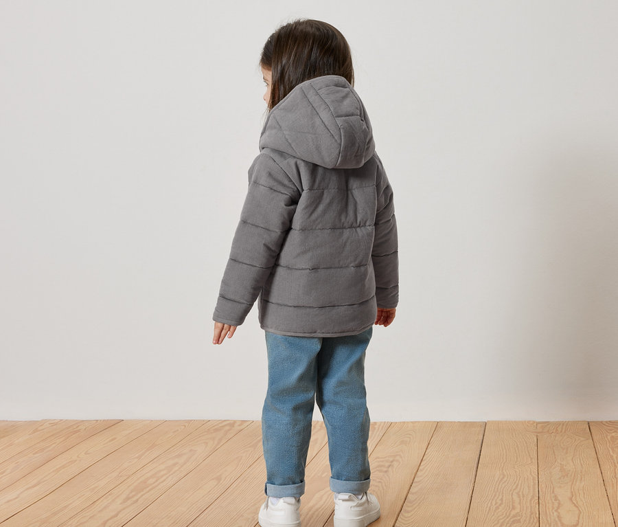 Kind von hinten, trägt eine graue Steppjacke mit Kapuze, Jeans und weiße Turnschuhe.