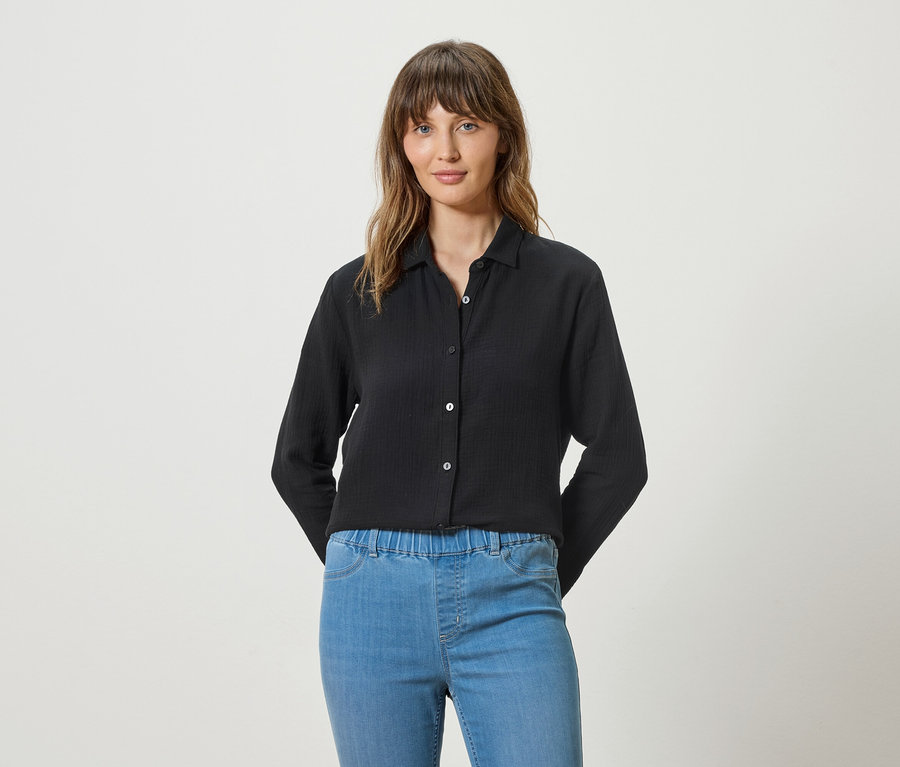 Frau trägt eine schwarze Langarmbluse und blaue Jeans vor einem weißen Hintergrund.
