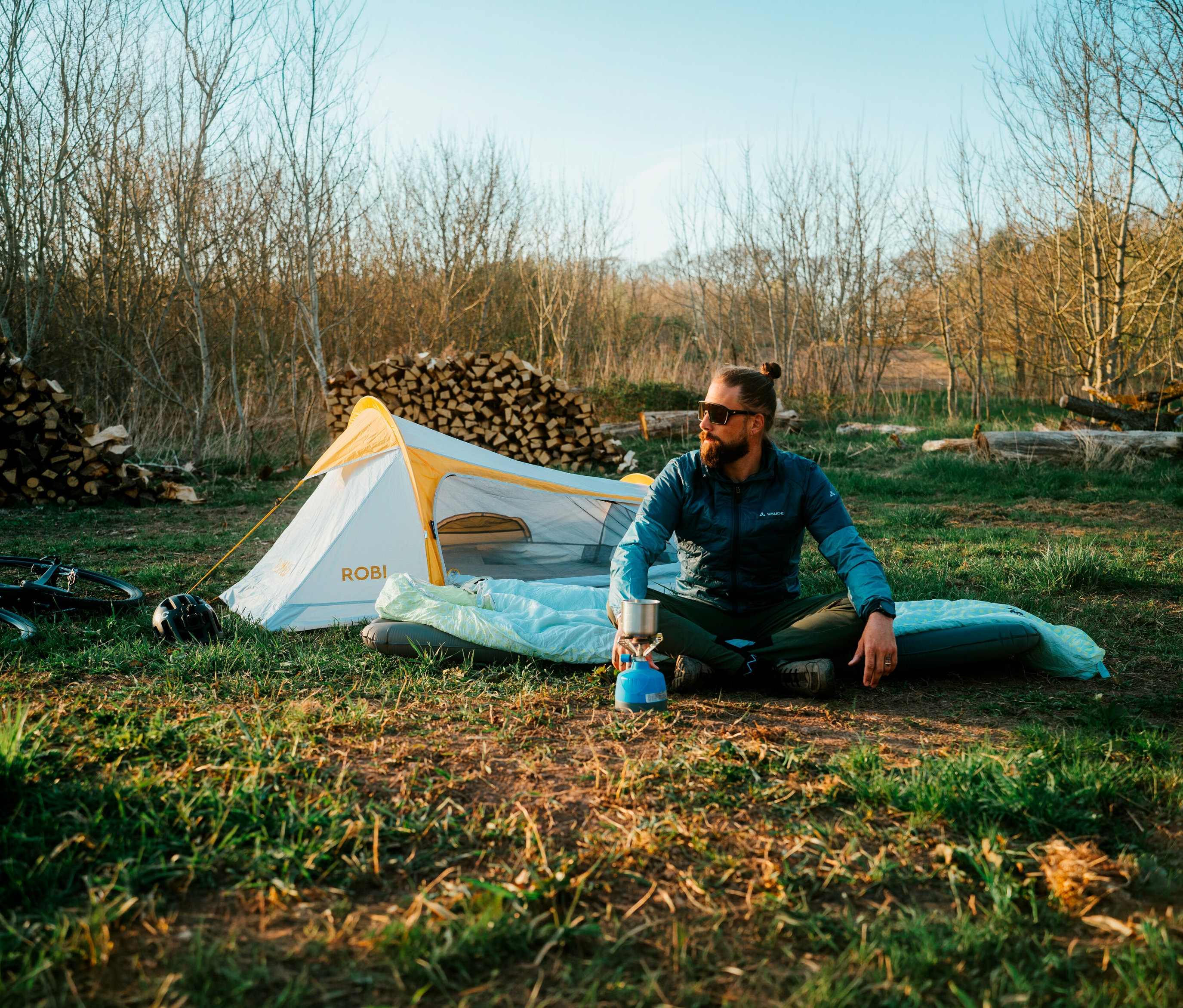 Mann sitzt vor Tambu »ROBI« 2-Personen-Leichtgewichtszelt auf einer Isomatte und kocht Kaffee. Im Hintergrund liegt ein Holzstapel.