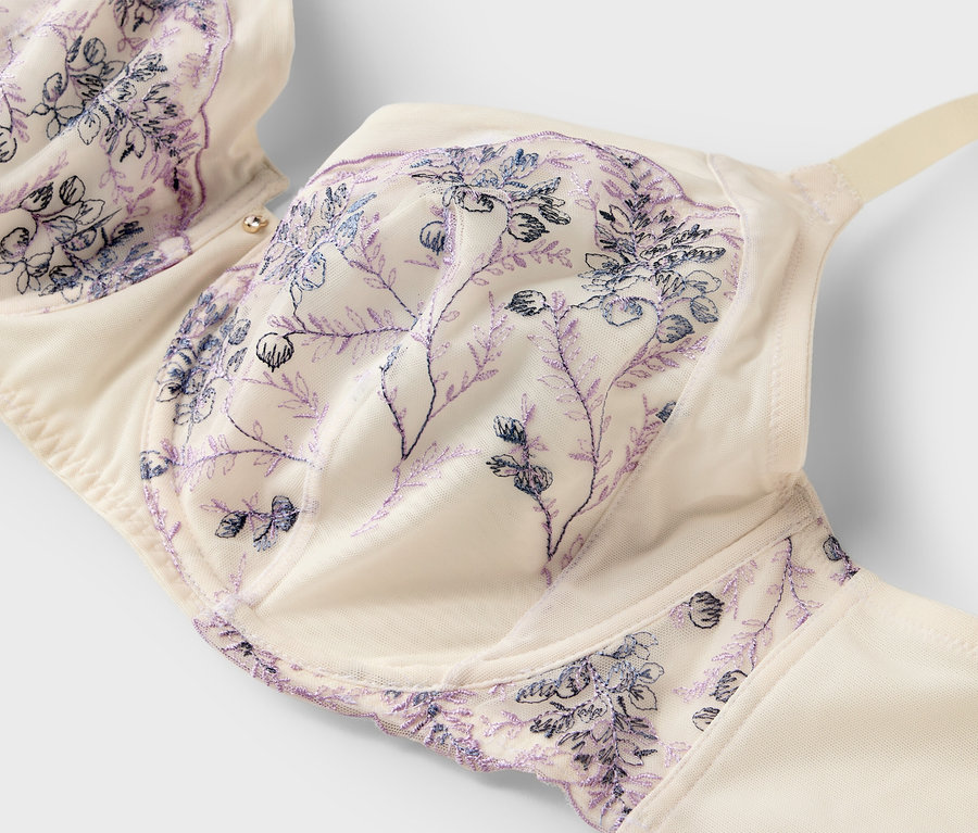 Nahaufnahme eines cremefarbenen BHs mit zarten lila Blumenstickereien auf weißem Hintergrund.