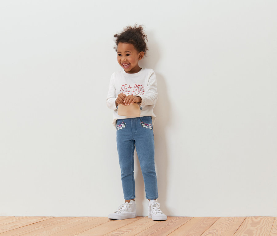 Lächelndes, lockiges Mädchen mit einer beigen Handtasche, einem langärmligen Oberteil und blauen Jeans mit Blumenstickerei, stehend vor einer weißen Wand.