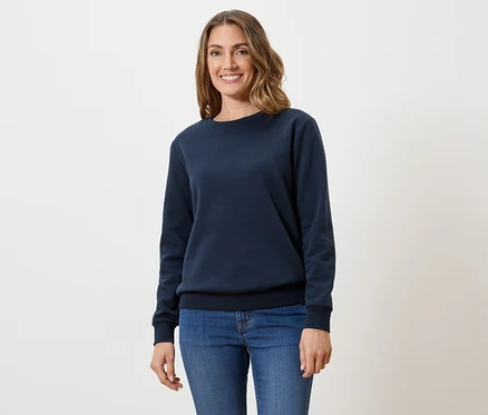 Eine Frau mit gewelltem, braunem Haar trägt einen marineblauen Pullover und Jeans vor einem weißen Hintergrund.