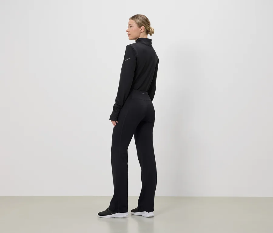Eine Frau steht seitlich in schwarzer Sportbekleidung, bestehend aus Oberteil, Hose und Schuhen, vor einem weißen Hintergrund.