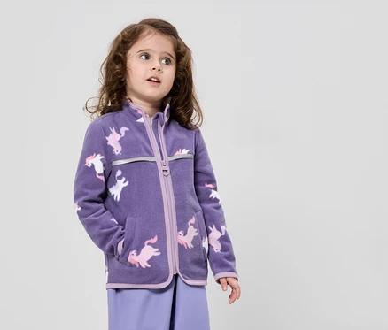 Mädchen mit braunen Locken trägt eine lila Fleecejacke mit Einhornmuster und lila Hose vor einem neutralen Hintergrund.