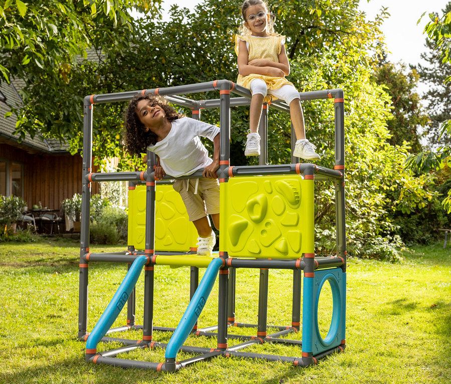 Zwei Kinder spielen auf einem Klettergerüst im Garten. Ein Kind sitzt oben, das andere lehnt sich an der Seite an.
