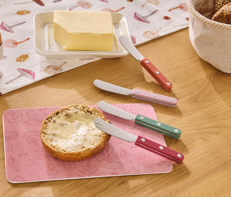 Auf einem Holztisch liegen ein Ei im Eierbecher, Butter, ein Brötchen mit Butter und vier Buttermesser in Rot, Rosa, Grün und Rot.