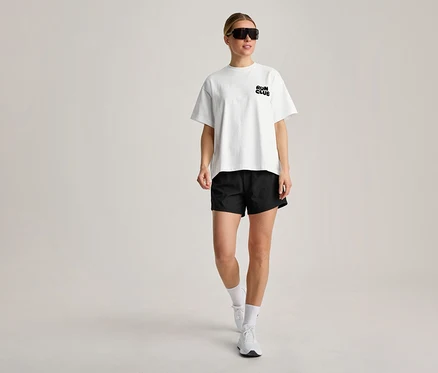 Eine Frau trägt ein weißes Sportshirt mit „RUN CLUB“-Aufschrift, schwarze Shorts, weiße Socken, weiße Turnschuhe und schwarze Sonnenbrille, während sie geht.
