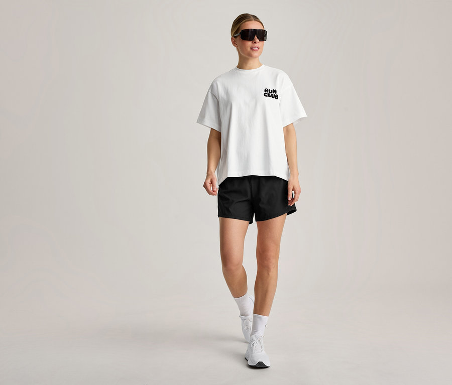 Eine Frau trägt ein weißes Sportshirt mit „RUN CLUB“-Aufschrift, schwarze Shorts, weiße Socken, weiße Turnschuhe und schwarze Sonnenbrille, während sie geht.
