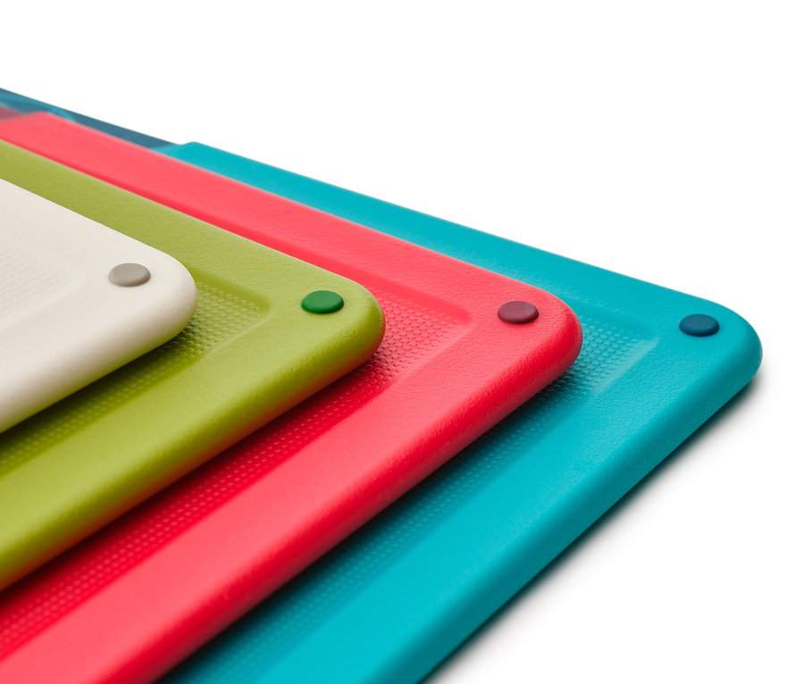 Nahaufnahme von vier bunten Schneidebrettern (weiß, grün, rot, türkis), die übereinander gestapelt sind und jeweils einen farblich passenden runden Gummistopfen in der Ecke haben.