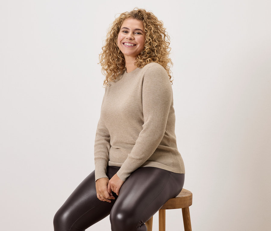 Frau mit lockigem Haar sitzt auf einem Holzschemel, trägt einen beigefarbenen Pullover und eine dunkle Leggings.