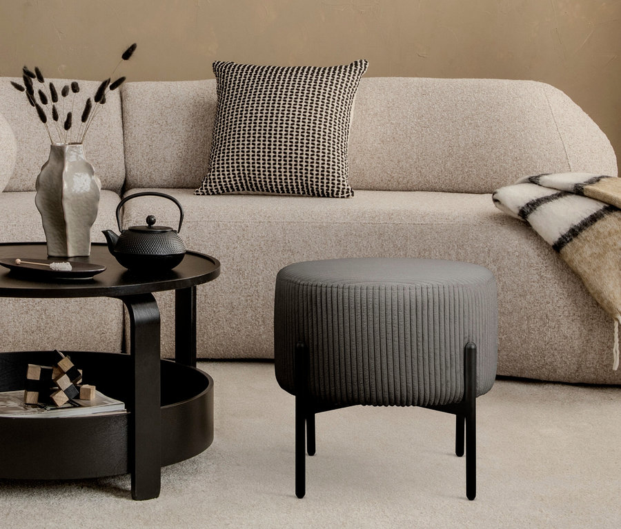 Ein grauer, gerippter Hocker steht vor einem beigen Sofa mit einem gemusterten Kissen. Eine schwarze Teekanne steht auf einem schwarzen Tisch.