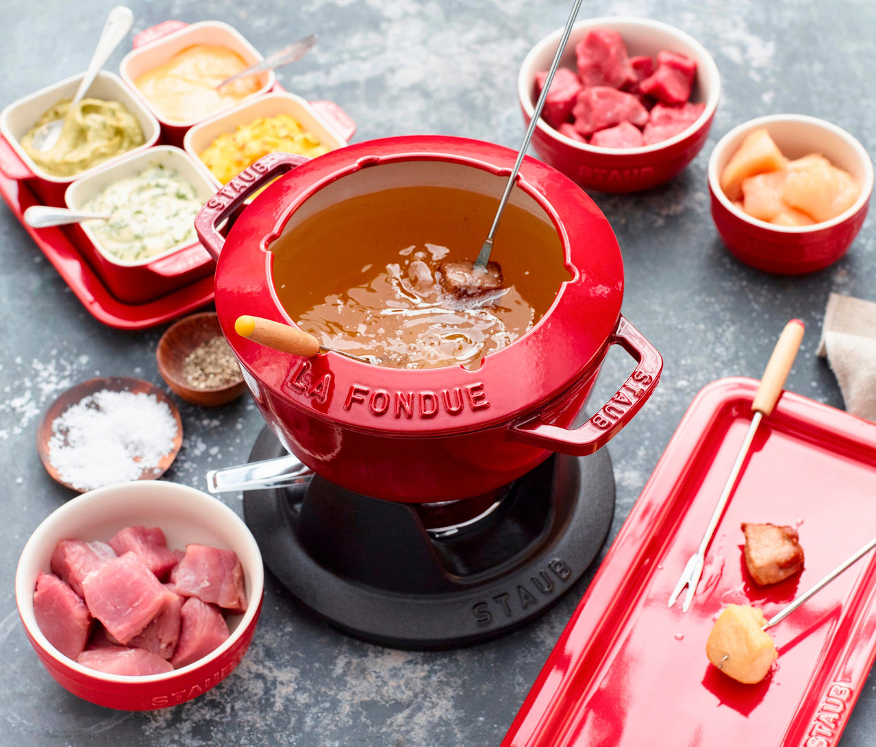 Ein rotes STAUB Fondue-Set steht auf einem Tisch, umgeben von Schüsseln mit Dips, Fleischwürfeln und Melonenspalten.