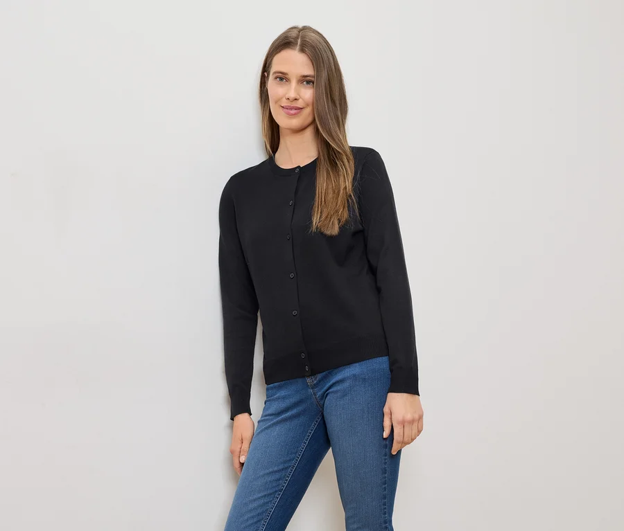 Frau mit langen braunen Haaren, schwarzer Strickjacke und blauen Jeans lehnt an einer weißen Wand.