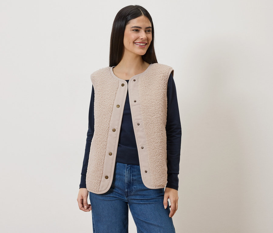 Frau mit langem, dunklem Haar trägt eine ärmellose, hellbraune Teddyfellweste über einem marineblauen Langarmshirt und Jeans.