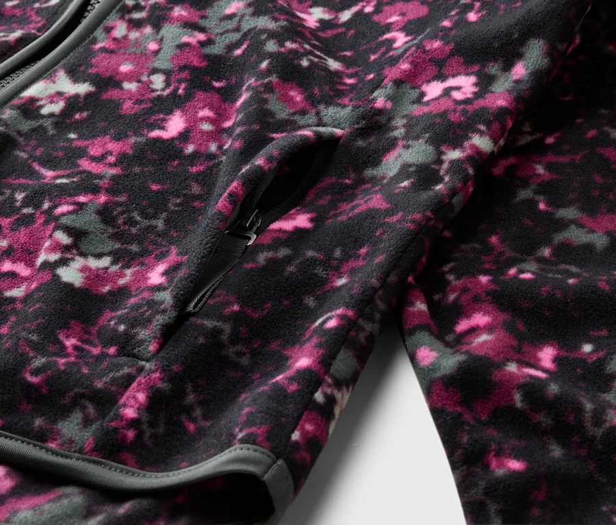Nahaufnahme einer Fleecejacke mit Reißverschluss und einem Muster aus Rosa, Grau und Schwarz. Eine Reißverschlusstasche ist sichtbar.
