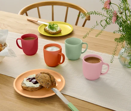 Holztisch mit hellem Tischläufer, vier bunten Tassen (rot, orange, mintgrün, rosa) mit Getränken, zwei Tellern mit belegten Brötchen, Brotkorb und Blumenvase.