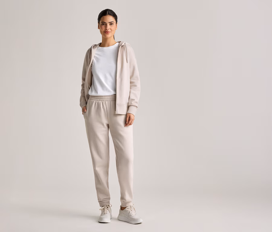 Frau mit Zopf trägt beiges Trainingsoutfit mit Kapuzenjacke, T-Shirt und Jogginghose, dazu weiße Turnschuhe.