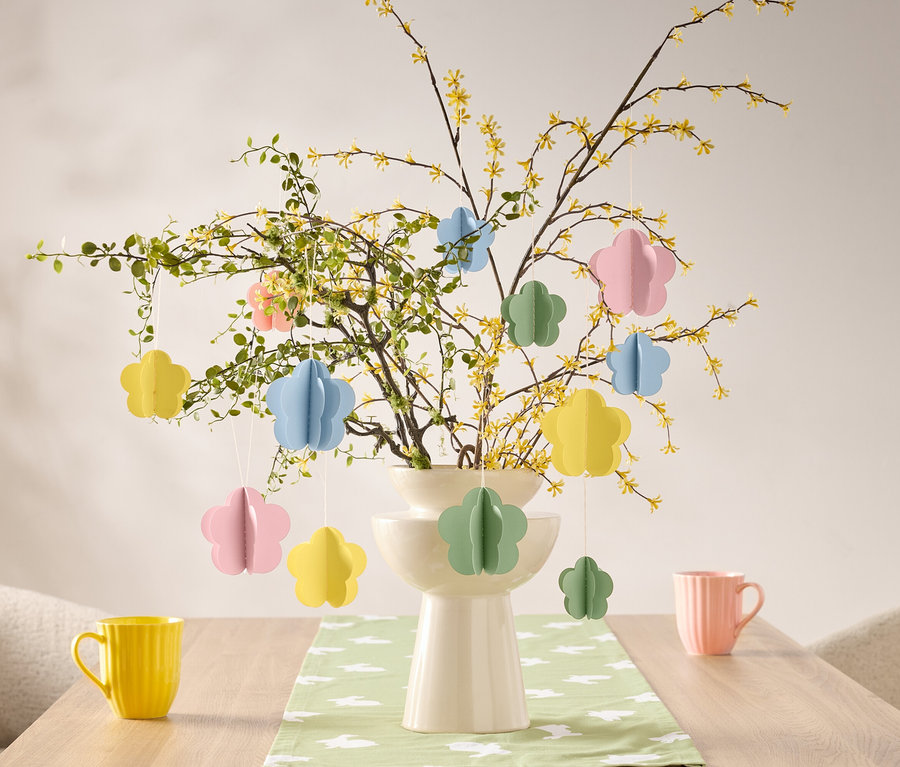 Blumenschmuck hängt an einem Zweig in einer Vase auf einem Tisch, neben einer gelben und einer rosa Tasse.