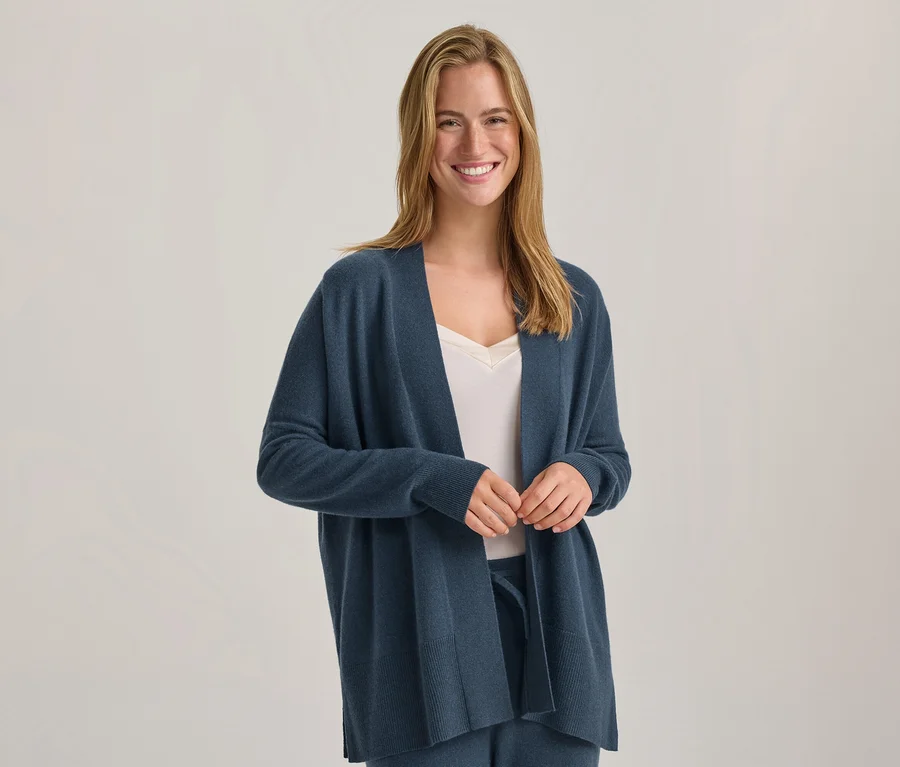 Lächelnde Frau mit langen blonden Haaren, die einen blauen offenen Cardigan über einem weißen Top und blauen Hosen trägt, die Hände gefaltet.