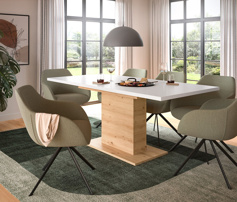 Ein modernes Esszimmer mit einem Esstisch mit weißer Platte und hellem Holzfuß, umgeben von sechs olivgrünen Stühlen mit schwarzen Metallbeinen. Ein grüner Teppich mit geometrischem Muster liegt auf dem Boden. Auf dem Tisch stehen eine Platte mit Snacks und Weingläser. Eine große Monstera-Pflanze steht neben einem gerahmten, abstrakten Kunstwerk. Große Fenster bieten Blick auf einen Garten, und eine graue Hängeleuchte hängt über dem Tisch.
