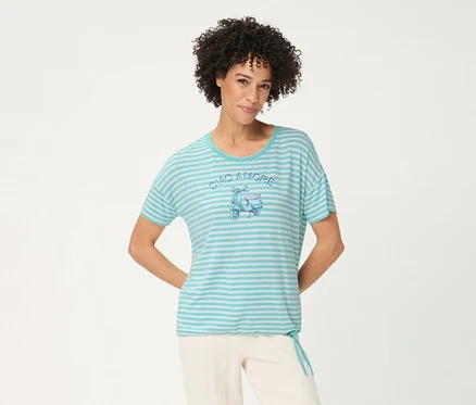 Lächelnde Frau mit lockigem Haar trägt ein gestreiftes Olsen T-Shirt in Türkis und Weiß mit einem 'CIAO AMORE' Roller-Motiv und heller Hose, die Hände hinter dem Rücken.