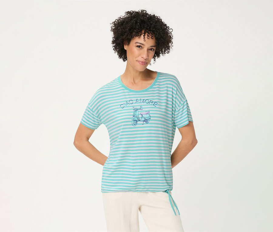 Lächelnde Frau mit lockigem Haar trägt ein gestreiftes Olsen T-Shirt in Türkis und Weiß mit einem 'CIAO AMORE' Roller-Motiv und heller Hose, die Hände hinter dem Rücken.