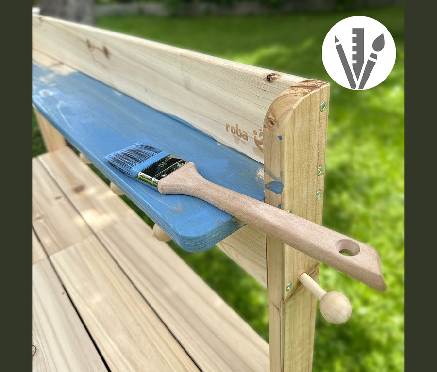 Holzbank wird blau gestrichen, ein Pinsel liegt auf der teilweise bemalten Oberfläche. Im Hintergrund ist grünes Gras zu sehen. Roba-Logo.