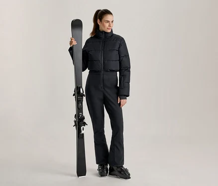 Frau in schwarzer, kurzer Daunen-Skijacke und schwarzer Skihose, die einen schwarzen Ski vertikal hält und schwarze Skischuhe trägt.