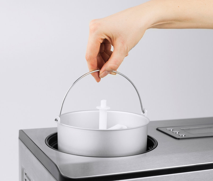 Eine Hand senkt einen silbernen Metalleimer mit einem weißen Plastikpaddel in eine Eismaschine aus Edelstahl.