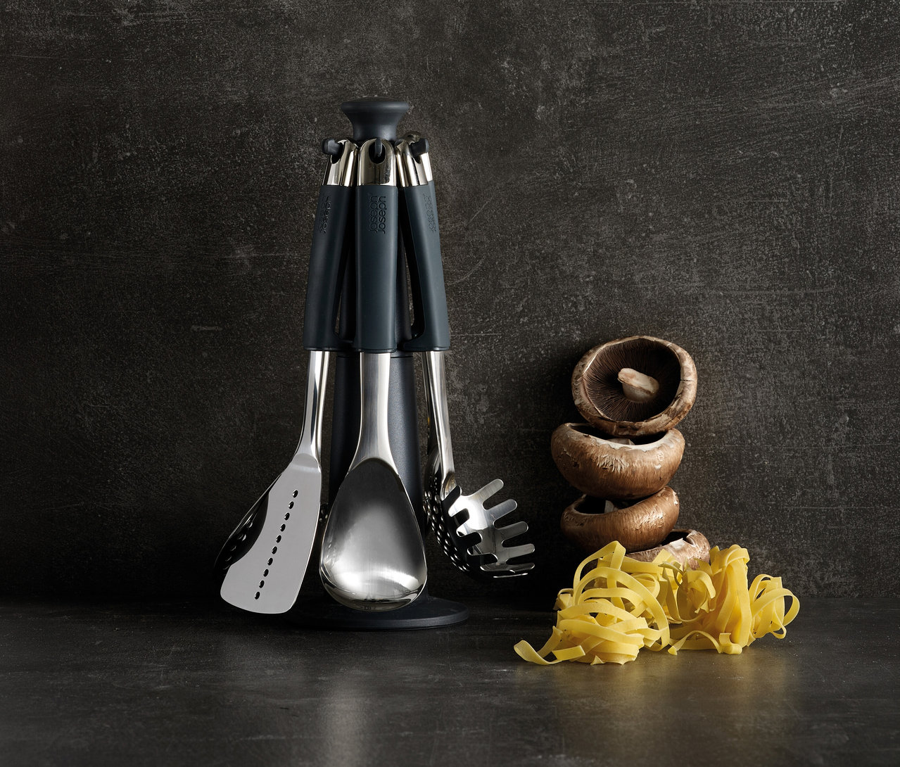 Joseph Joseph Elevate Steel Carousel mit Edelstahl-Küchenutensilien (Pfannenwender, Löffel, Pastalöffel) auf dunkler Oberfläche. Rechts davon gestapelte Pilze und Tagliatelle-Nudeln.