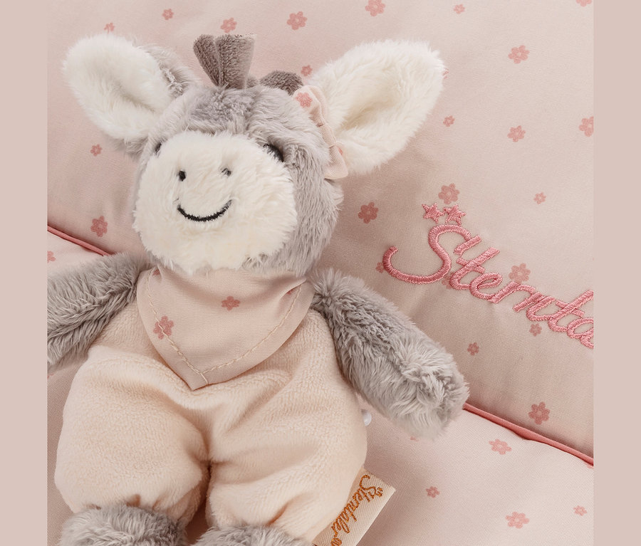 Graues und weißes Plüsch-Eselchen »Emmi Girl« von Sterntaler mit rosa Latzhose und Halstuch auf rosa Stoff mit Blumenmuster.