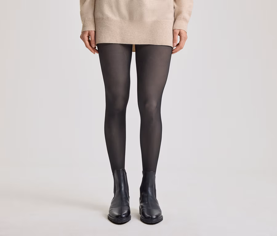 Eine Person steht in einem beigen Pullover, schwarzen Strumpfhosen und schwarzen Stiefeletten.