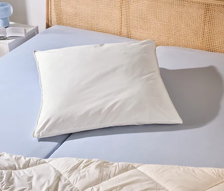 Ein Bett mit hellblauem Spannbettlaken, einem weißen Kissen, einer beigen Steppdecke und einem Rattan-Kopfteil. Links ein weißer Nachttisch mit Buch und blauer Lampe.