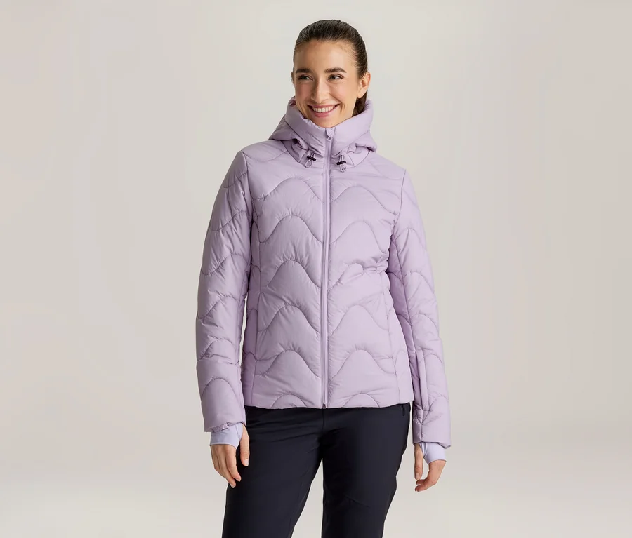 Lächelnde Frau mit zurückgebundenen dunklen Haaren, die eine hellviolette Steppjacke mit Wellenmuster und Kapuze sowie eine dunkle Hose trägt und nach rechts blickt.