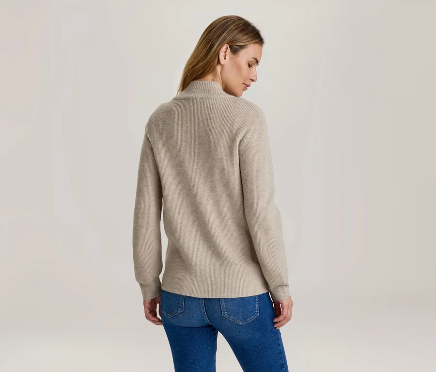 Frau von hinten, leicht gedreht, trägt einen beigen gerippten Rollkragenpullover und blaue Jeans.