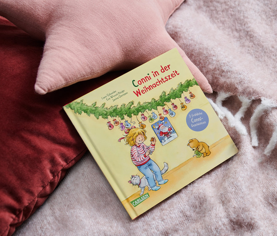 Das Buch Conni in der Weihnachtszeit liegt auf einer Decke neben rosa Kissen.
