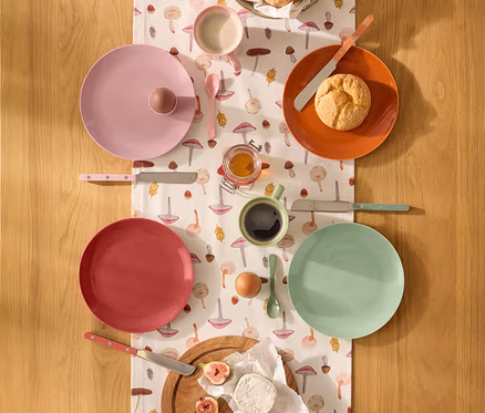 Blick von oben auf einen Holztisch mit einem Tischläufer mit Pilzmuster, der mit Frühstücksgeschirr und Speisen gedeckt ist. Es gibt vier Teller in Rosa, Orange, Rot und Mintgrün, eine Schale mit Brötchen, Honig, ein Ei, Feigen und Käse. Dazu passendes Besteck und zwei Tassen sind ebenfalls zu sehen.