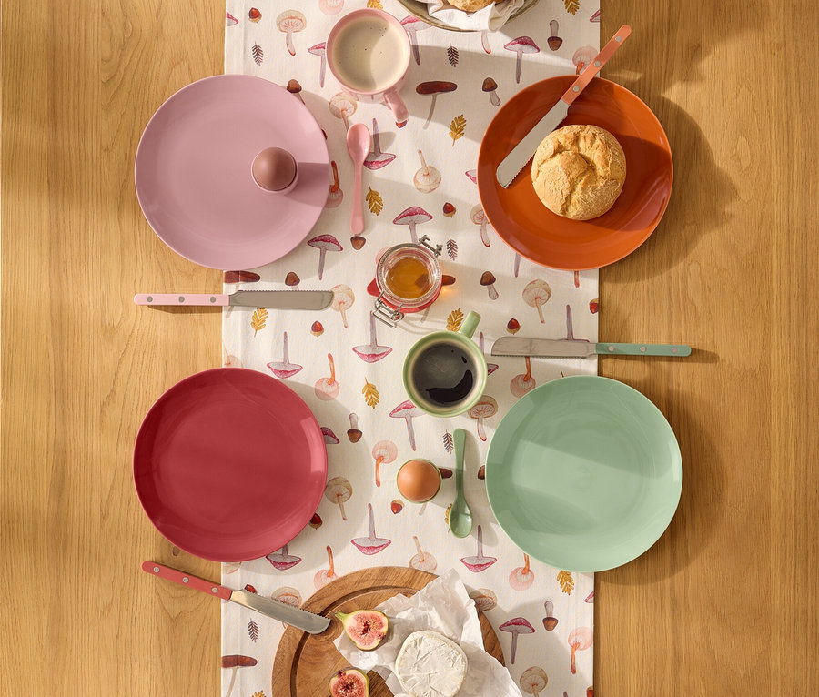 Blick von oben auf einen Holztisch mit einem Tischläufer mit Pilzmuster, der mit Frühstücksgeschirr und Speisen gedeckt ist. Es gibt vier Teller in Rosa, Orange, Rot und Mintgrün, eine Schale mit Brötchen, Honig, ein Ei, Feigen und Käse. Dazu passendes Besteck und zwei Tassen sind ebenfalls zu sehen.