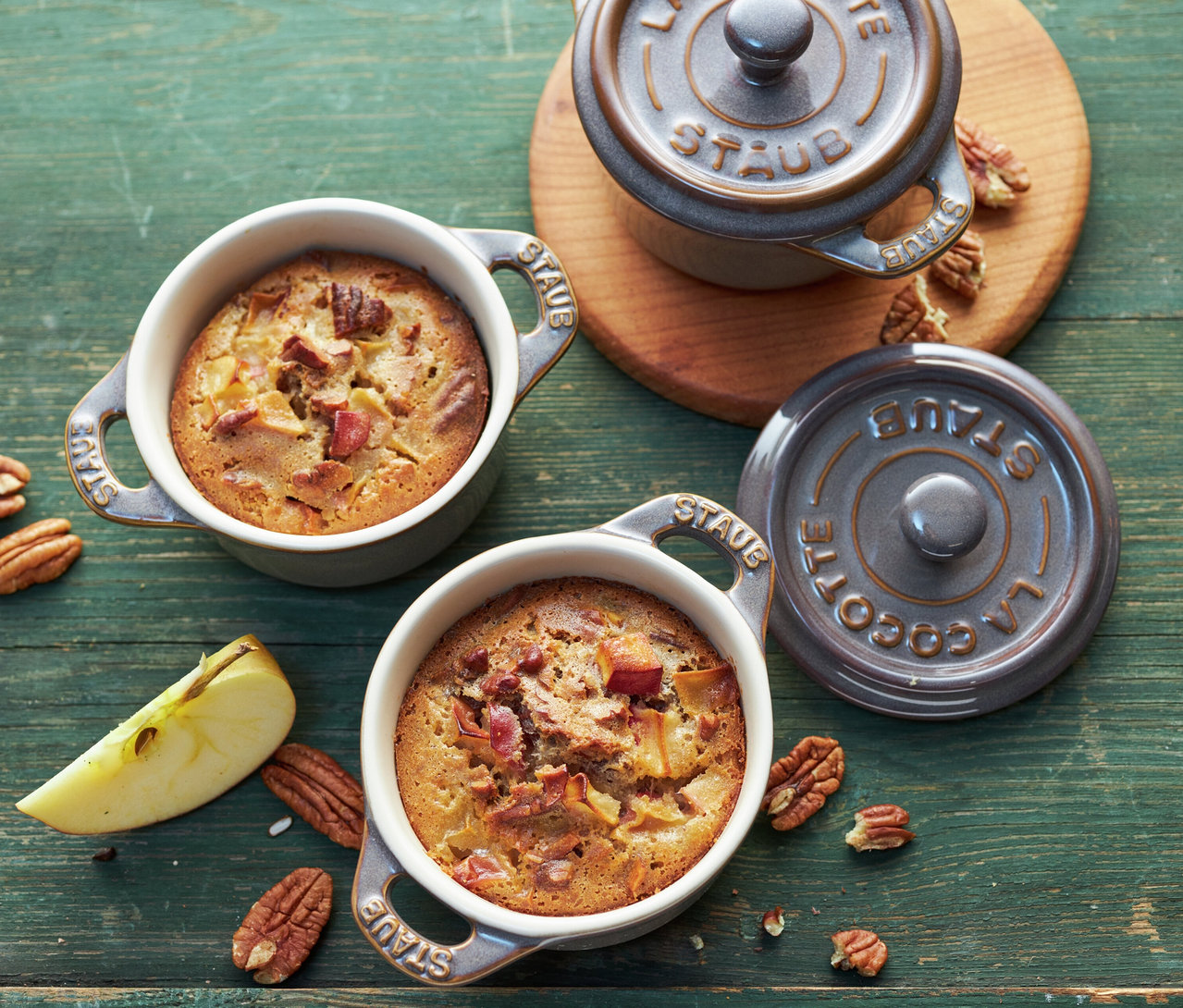 Drei der 4 STAUB Keramik-Cocottes in Antikgrau, gefüllt mit Apfel-Pekannuss-Dessert, stehen auf einem grünen Holztisch, umgeben von Nüssen und Apfelstücken.