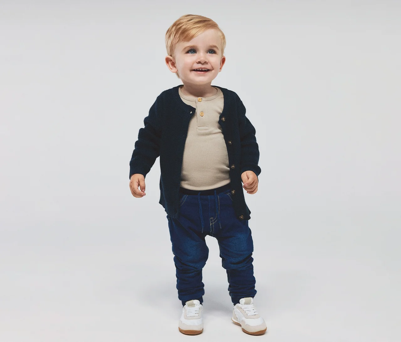 Ein lächelnder Kleinkindjunge mit blonden Haaren und blauen Augen, bekleidet mit einem beigen Langarmshirt, einem marineblauen Strickcardigan, dunkelblauen Jeans und weißen und beigen Turnschuhen.