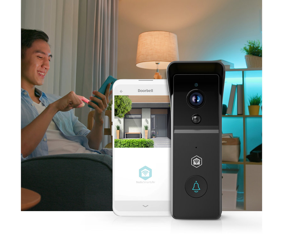 Ein lächelnder Mann sitzt auf einem Sofa und bedient ein Smartphone, das eine Türklingel-App mit einem Hauseingang anzeigt. Daneben steht eine schwarze Smart-Türklingel.