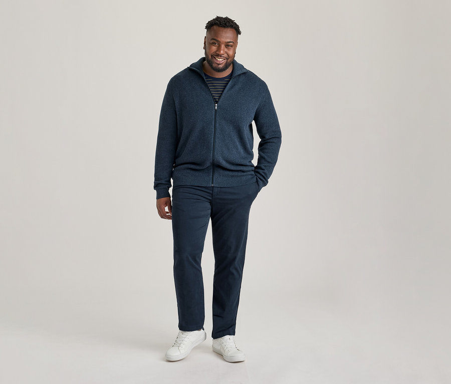 Mann trägt einen marineblauen Reißverschluss-Cardigan über einem gestreiften Shirt und eine marineblaue Chino, lächelt.