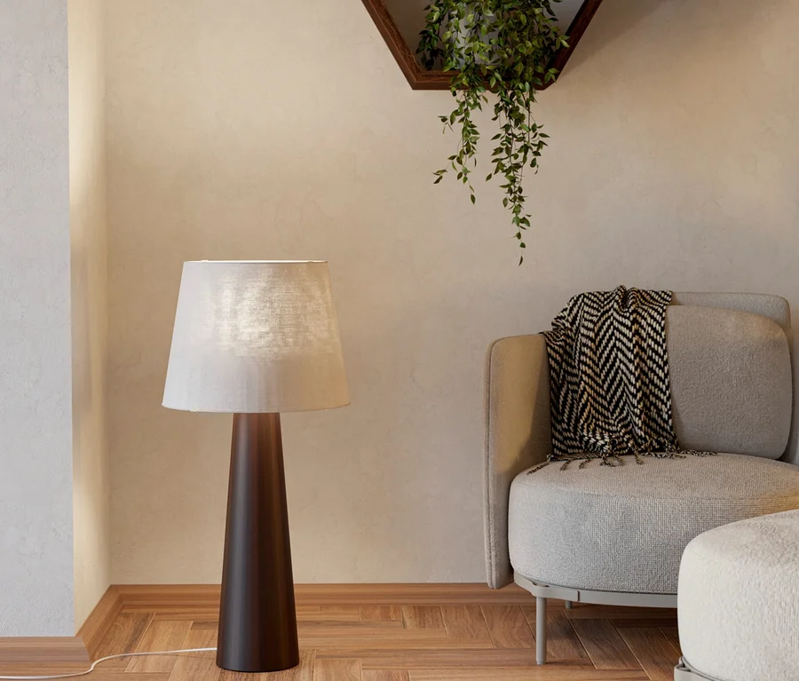 Eine Stehlampe mit dunklem Fuß und hellem Schirm steht neben einem hellgrauen Sessel mit gemusterter Decke und passendem Hocker. Ein heller Teppich liegt auf einem Fischgrätparkettboden, und zwei sechseckige Holzregale an der Wand zeigen eine Hängepflanze und eine Vase mit einer Trockenblume.