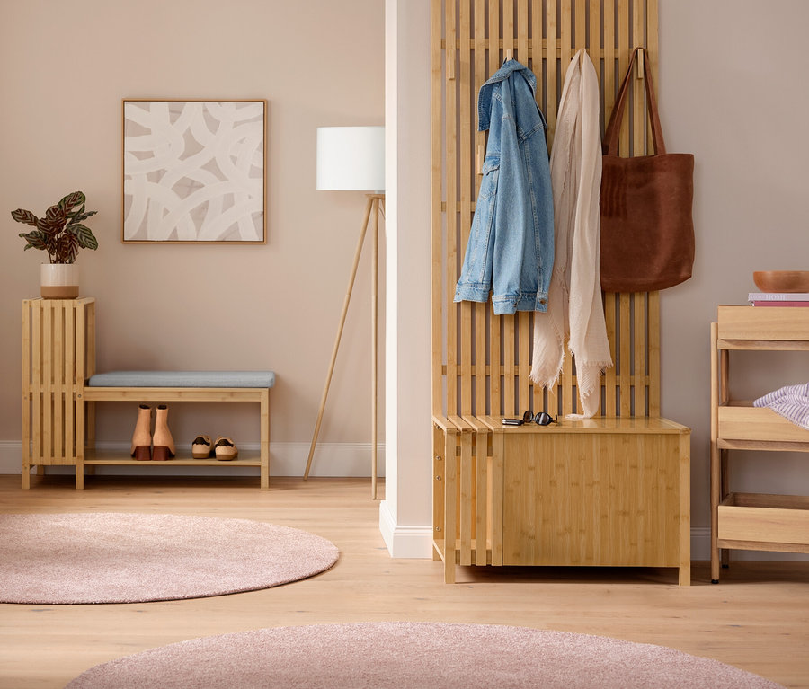 Heller Vorraum mit einem Kleiderständer mit Ablage, an dem Jacken hängen. In der Mitte eine Sitzbank mit Schuhfach und Ablage in Lamellenoptik mit Kissen und Schuhen darunter, daneben eine Dreibein-Stehleuchte aus Holz. Rechts eine Garderobe mit Sitzbank in Lamellenoptik mit Jacke, Schal und Tasche. Daneben ein Standregal aus Massivholz mit 3 Fächern. Auf dem Boden liegen zwei pinke Teppiche „Charme“.