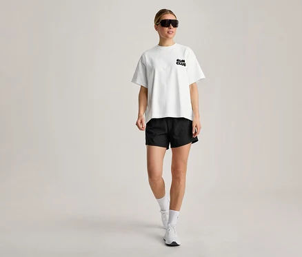 Eine Frau trägt ein weißes Sportshirt mit „RUN CLUB“-Aufschrift, schwarze Shorts, weiße Socken, weiße Turnschuhe und schwarze Sonnenbrille, während sie geht.