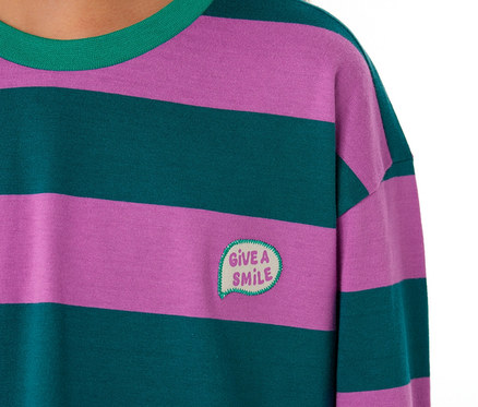 Nahaufnahme eines Kindes, das ein SCHIESSER Kinder-Pyjama Oberteil mit lila und dunkelgrünen Querstreifen, grünem Kragen und einem aufgestickten „GIVE A SMILE“-Sprechblasen-Patch trägt.