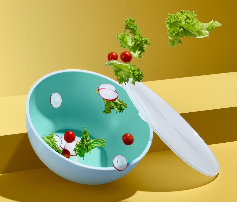 Grüner Salat und Radieschen fallen in eine Salatschüssel mit Deckel vor gelbem Hintergrund.