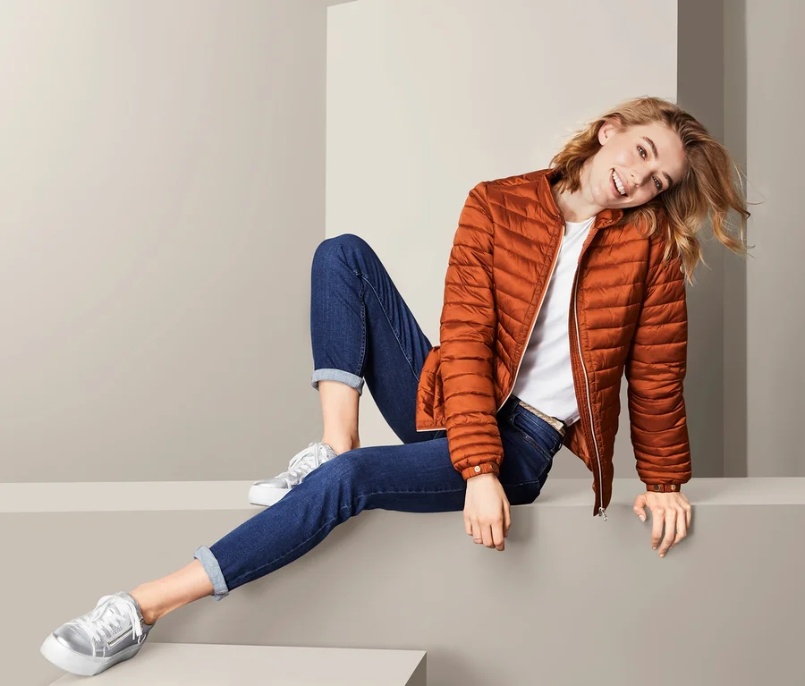 Frau sitzt und trägt eine Steppjacke, Jeans und Turnschuhe.