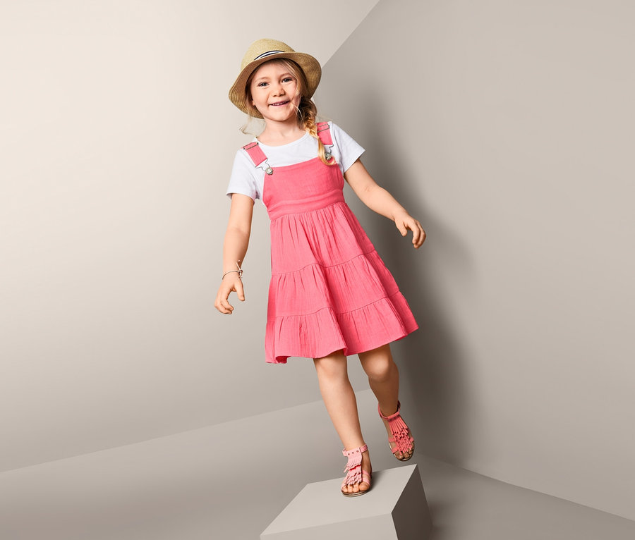Ein Mädchen steht auf einem Würfel und trägt ein Trägerkleid und einen Hut.